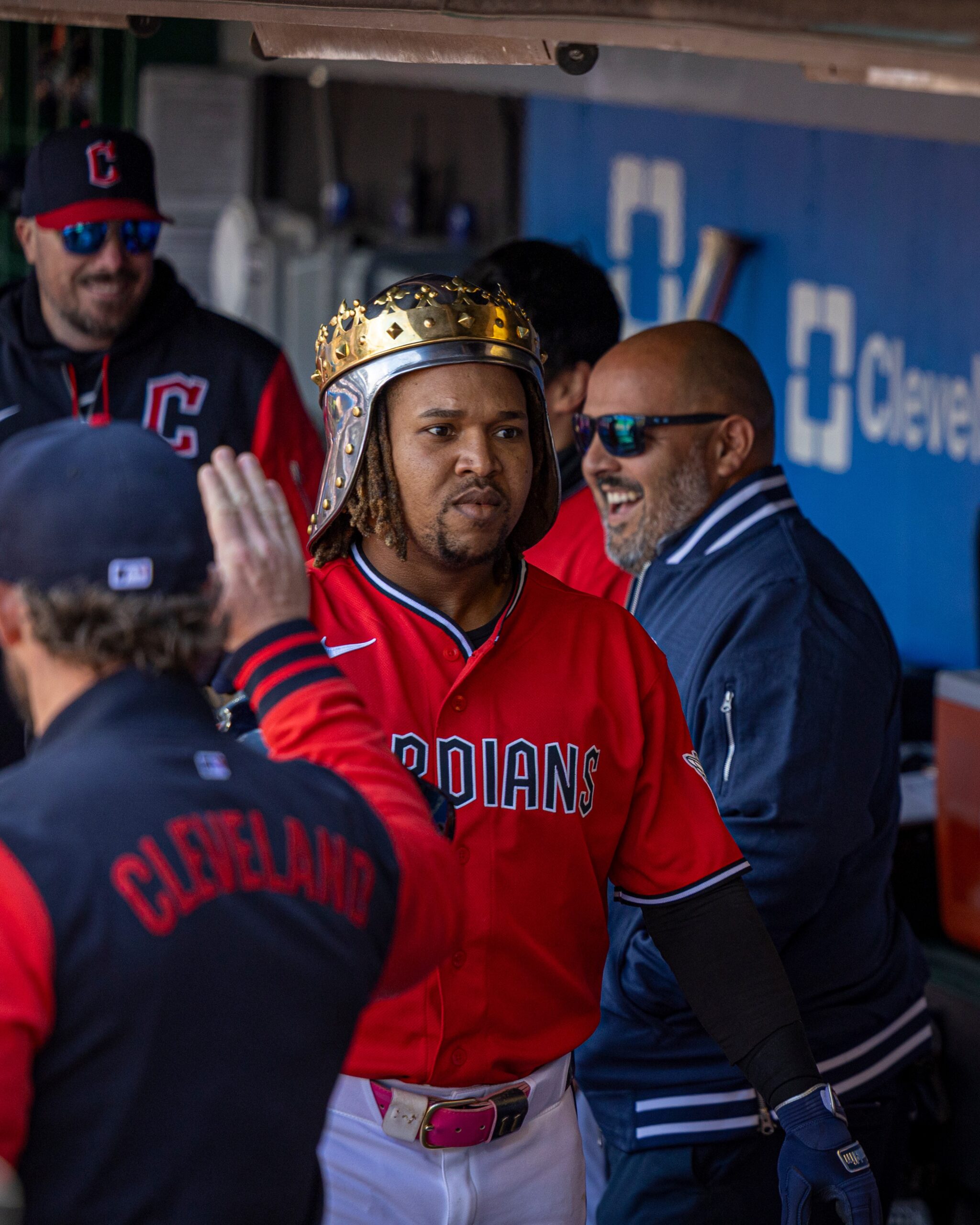Baseball player in a red Indians jersey wearing a gold crown helmet in the dugout, high-fiving a teammate.