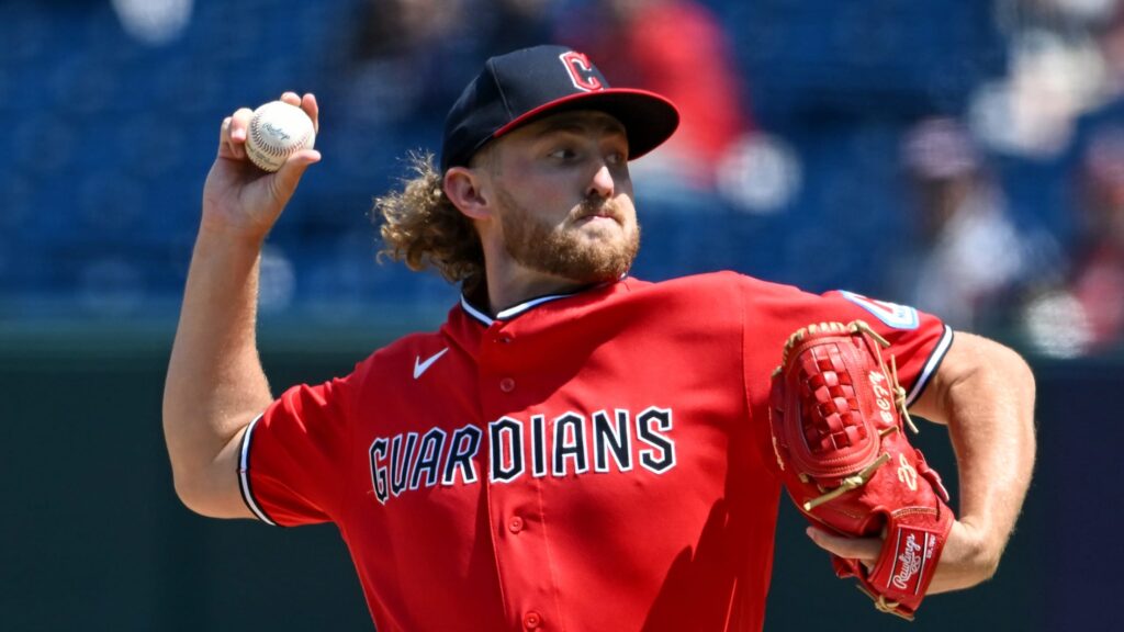 Guardians pitcher in a red uniform winds up to throw a baseball, with a red glove on his other hand against a blurred stadium background.