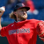 Guardians pitcher in a red uniform winds up to throw a baseball, with a red glove on his other hand against a blurred stadium background.