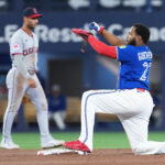 Blue-uniform baserunner slides into a base, reaching the bag with his left foot as a gray-uniform fielder watches in the background.