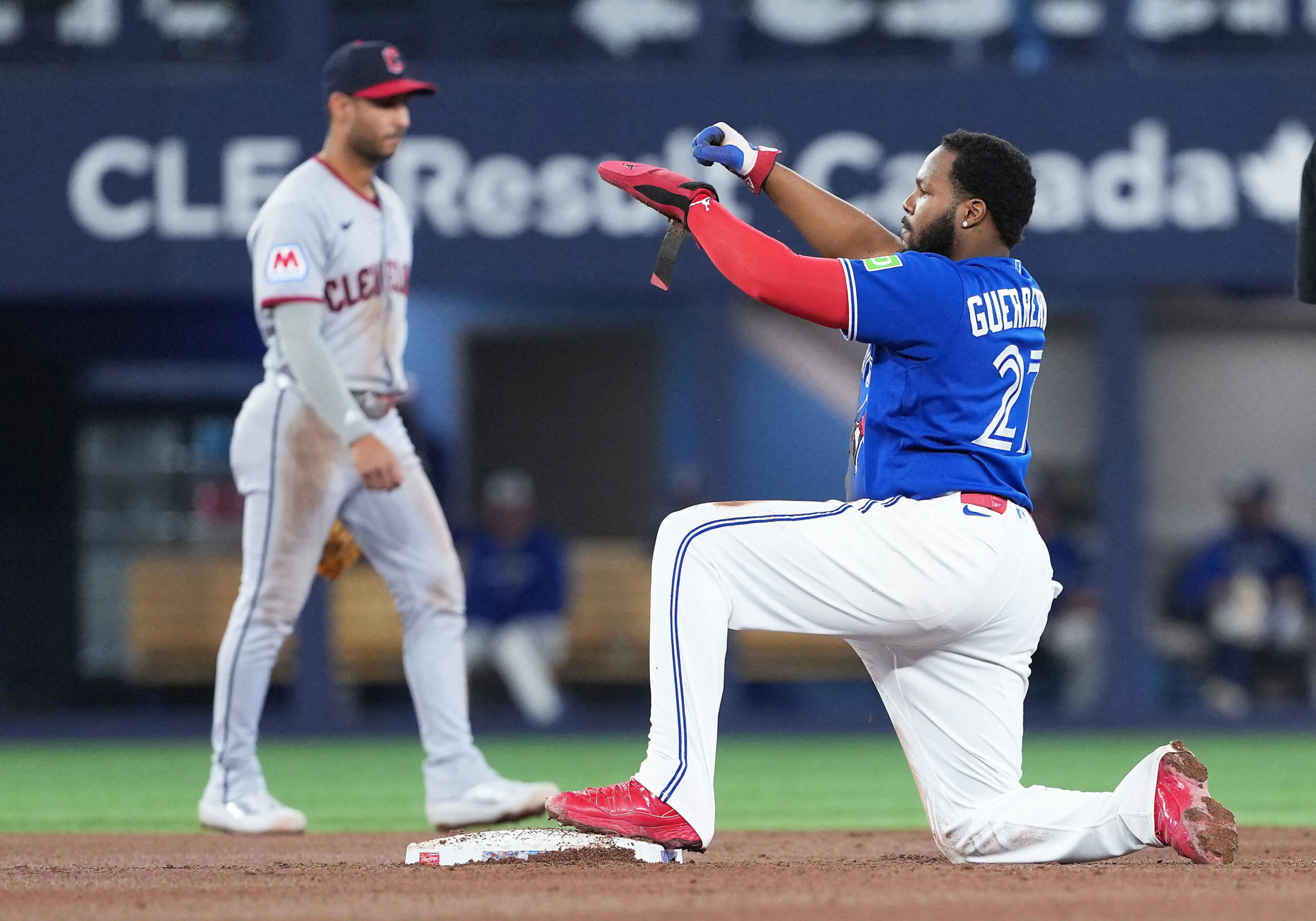 Blue-uniform baserunner slides into a base, reaching the bag with his left foot as a gray-uniform fielder watches in the background.