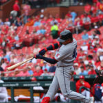 Baseball batter in gray swings at a pitch with the catcher in red gear behind him and a sea of red-clad fans in the stands behind.