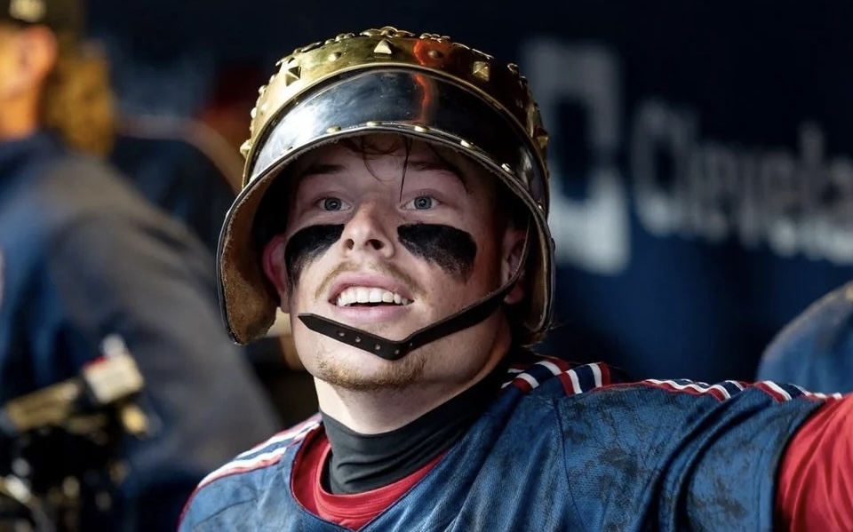 Young man wearing a firefighter helmet with black eye paint, smiling in a blue-red jersey indoors.