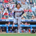 Cleveland baseball player sprinting on the field after a play, bat lying on the ground nearby with spectators in the stands behind him (outfield dugout visible).