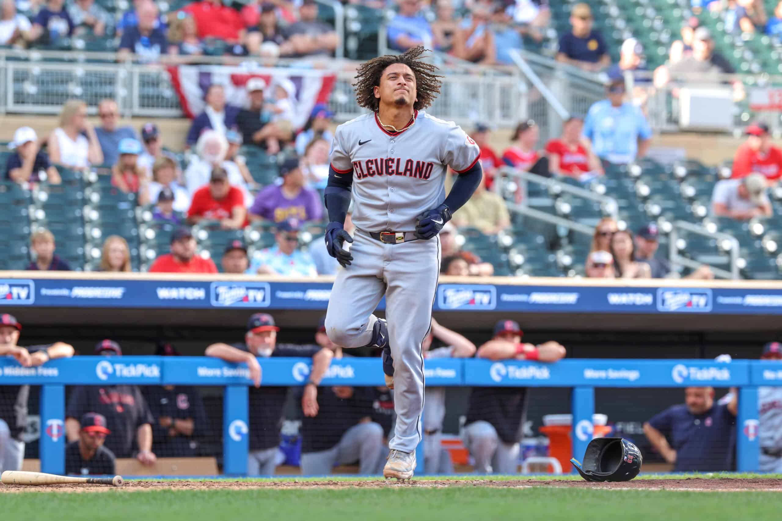 Cleveland baseball player sprinting on the field after a play, bat lying on the ground nearby with spectators in the stands behind him (outfield dugout visible).