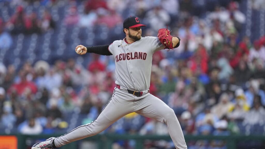 Cleveland pitcher in a gray uniform delivering a pitch on the mound with a crowd behind him.