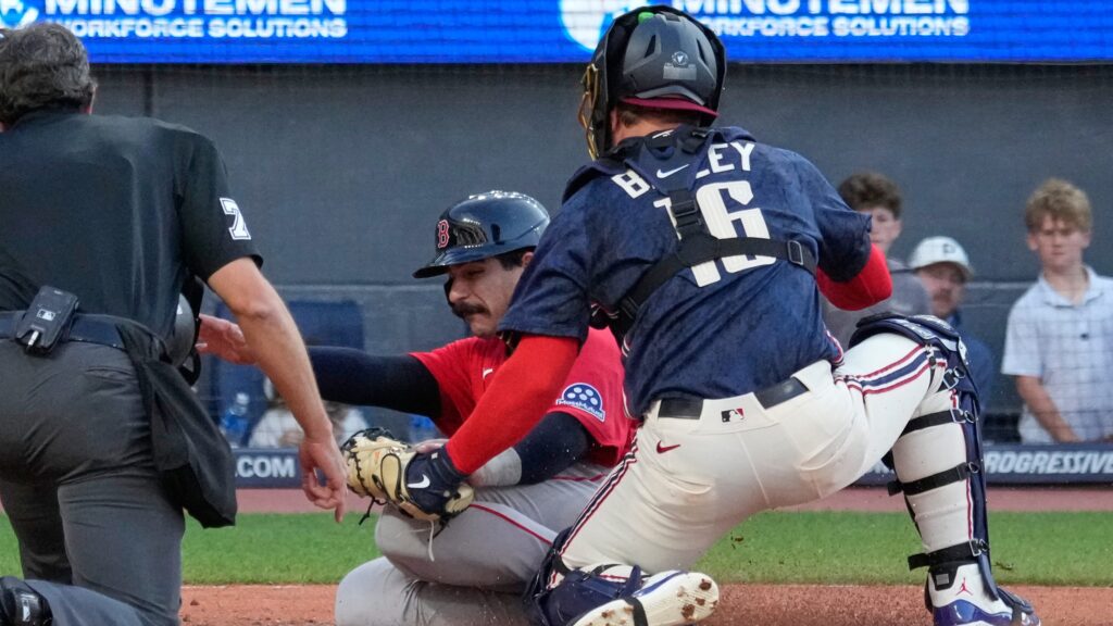 Baseball catcher in blue gear blocks a play at home plate as a runner in red slides in, with an umpire nearby watching the collision.