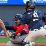 Baseball catcher in blue gear blocks a play at home plate as a runner in red slides in, with an umpire nearby watching the collision.
