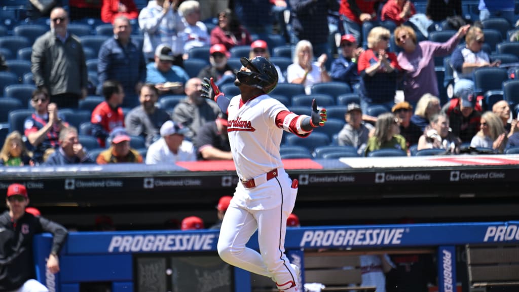 Baseball player in a white uniform with red trim raises arms as he runs, celebrating on the field with fans watching from the stands behind him.