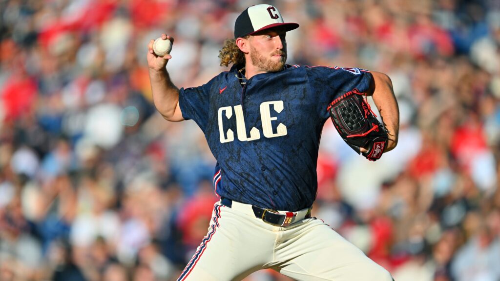 Baseball pitcher in a blue CLE jersey throws a ball with a glove on his right hand, crowd blurred in the stands behind him
