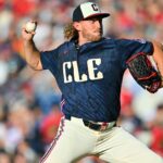 Baseball pitcher in a blue CLE jersey throws a ball with a glove on his right hand, crowd blurred in the stands behind him
