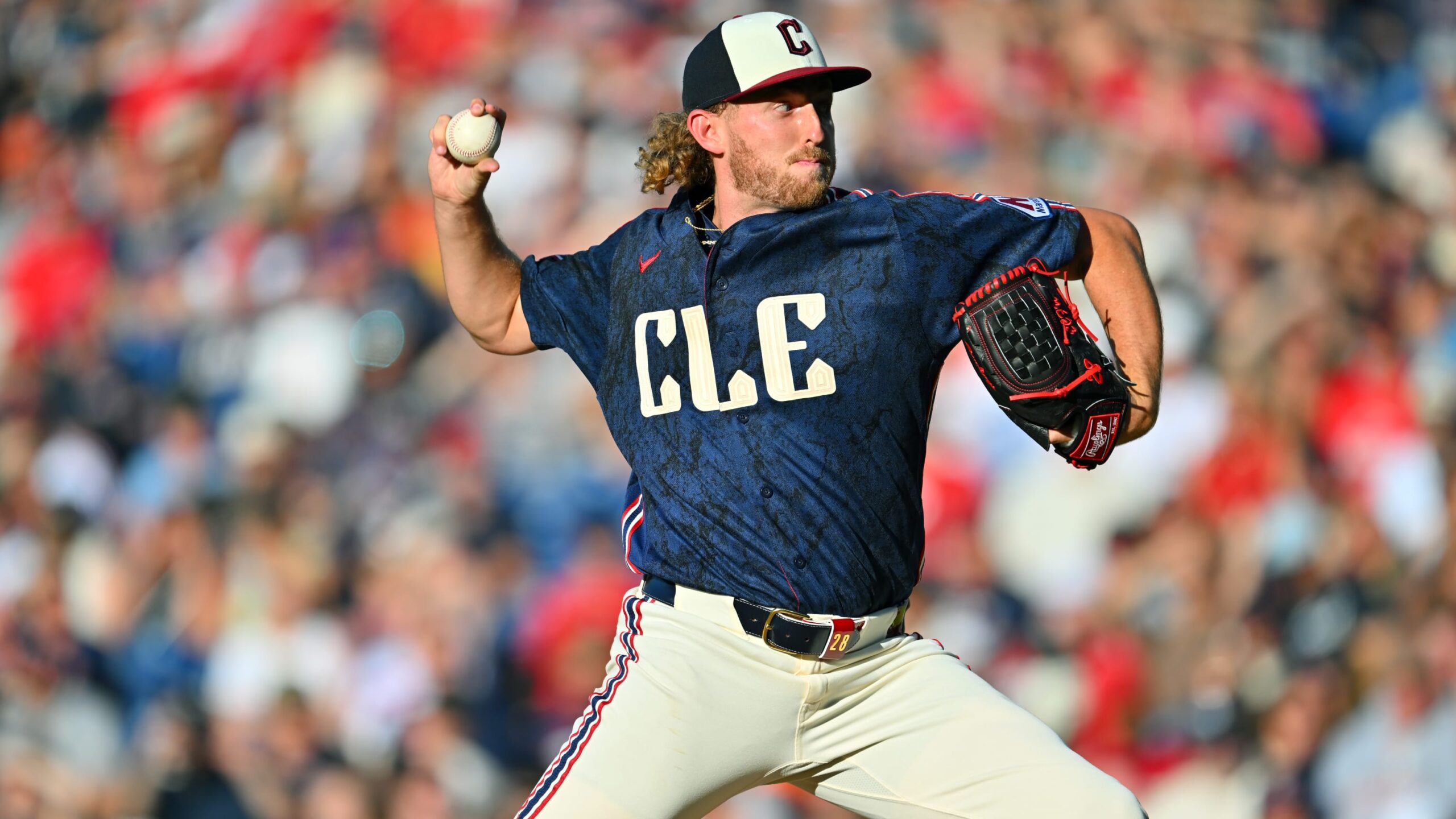 Baseball pitcher in a blue CLE jersey throws a ball with a glove on his right hand, crowd blurred in the stands behind him