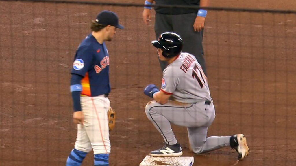 Baseball runner in gray slides into a white base as an umpire and a blue-uniform defender observe on a dirt infield.