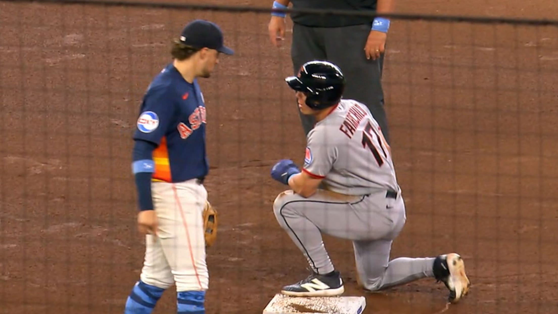 Baseball runner in gray slides into a white base as an umpire and a blue-uniform defender observe on a dirt infield.