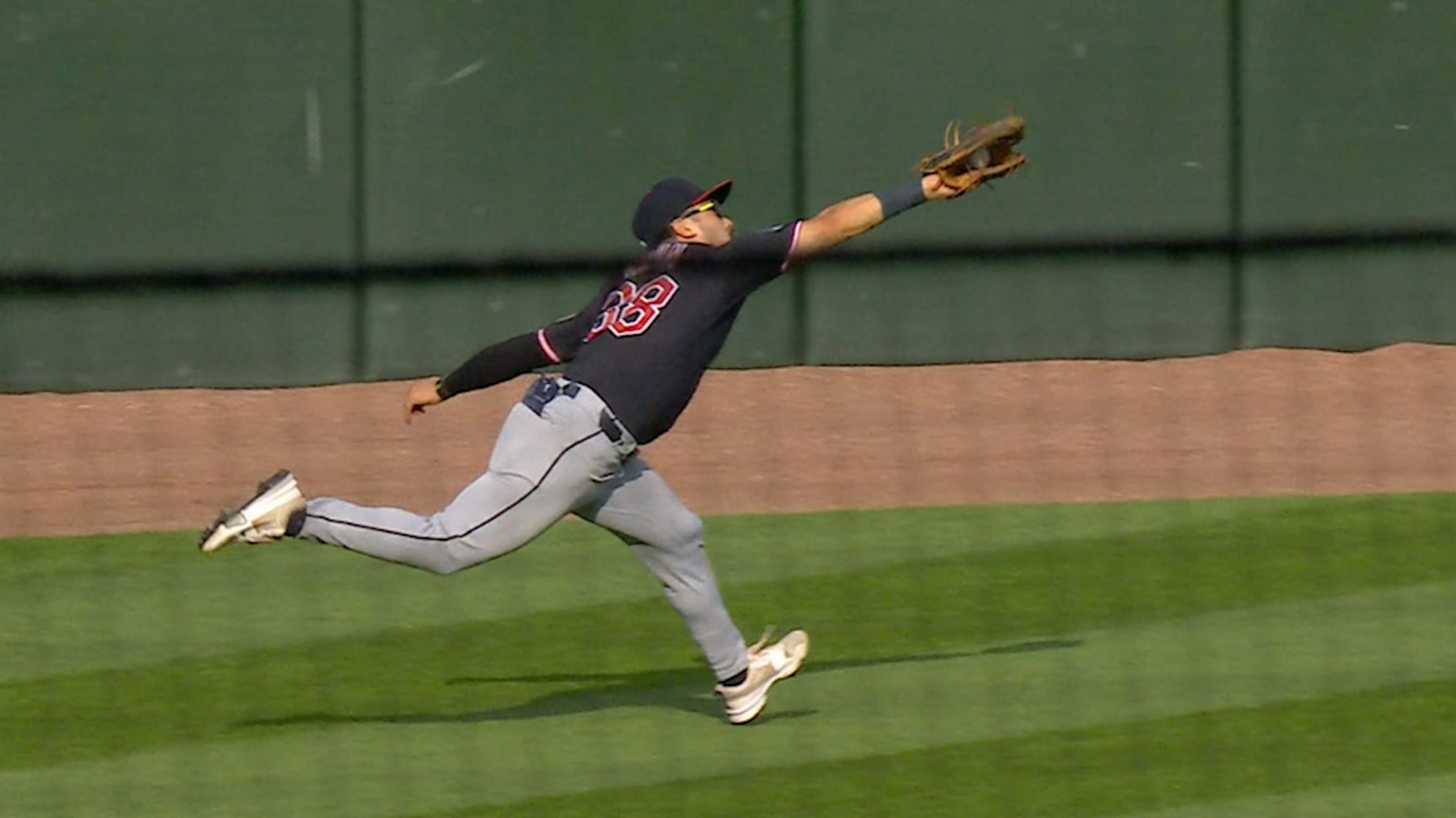 Baseball fielder in a dark uniform lunges forward with glove extended to catch a ball on the outfield grass.