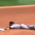 Baseball player slides head-first into a base on an orange dirt infield, reaching for the bag as a nearby runner moves in.