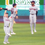Cleveland baseball players celebrate mid-air on the field, two jumping in the foreground with a smiling teammate nearby.