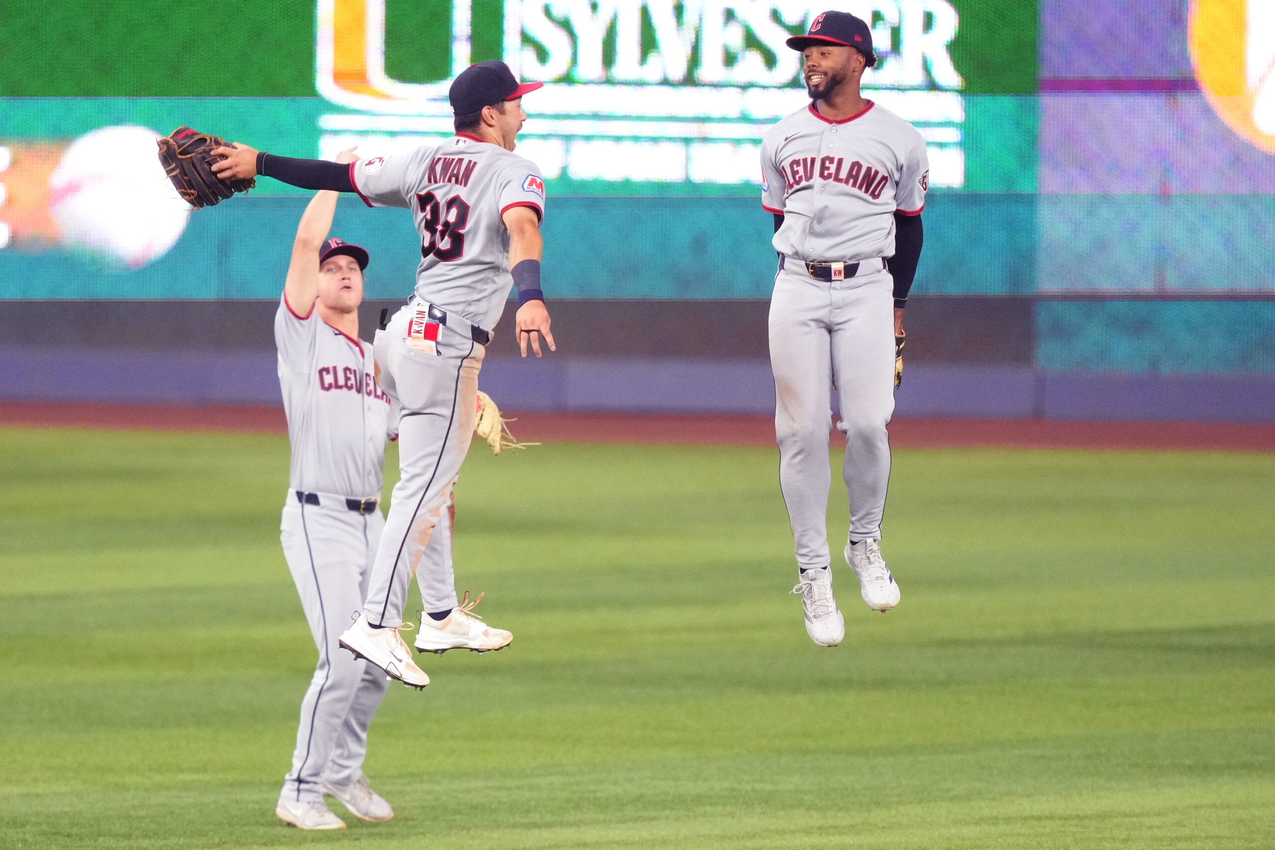 Cleveland baseball players celebrate mid-air on the field, two jumping in the foreground with a smiling teammate nearby.