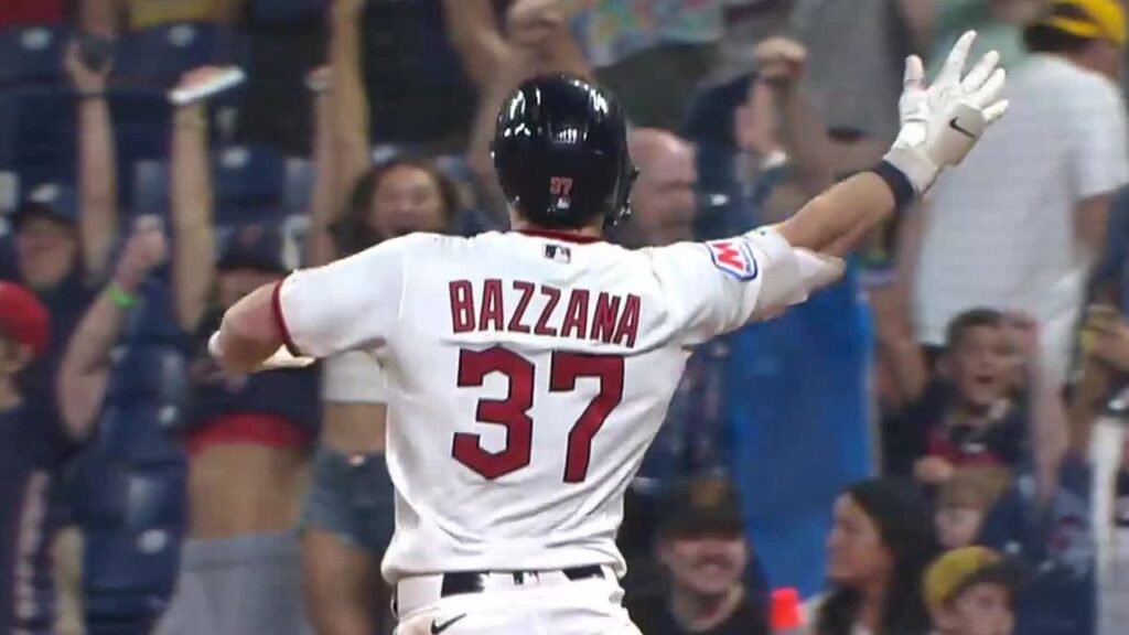Baseball player (back view) in a white uniform with 'BAZZANA' and number 37 raises arms in celebration as cheering fans look on from the stands.