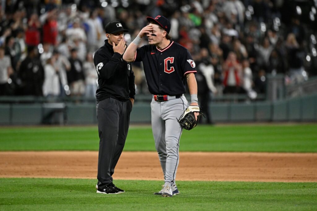 Baseball player in black uniform with a red 'C' on chest stands with an umpire on the infield; player shields his eyes with his hand while crowd watches.