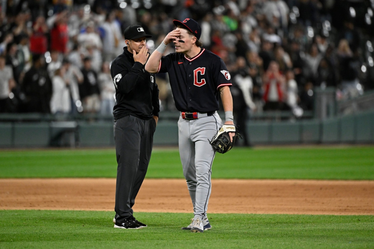 Baseball player in black uniform with a red 'C' on chest stands with an umpire on the infield; player shields his eyes with his hand while crowd watches.