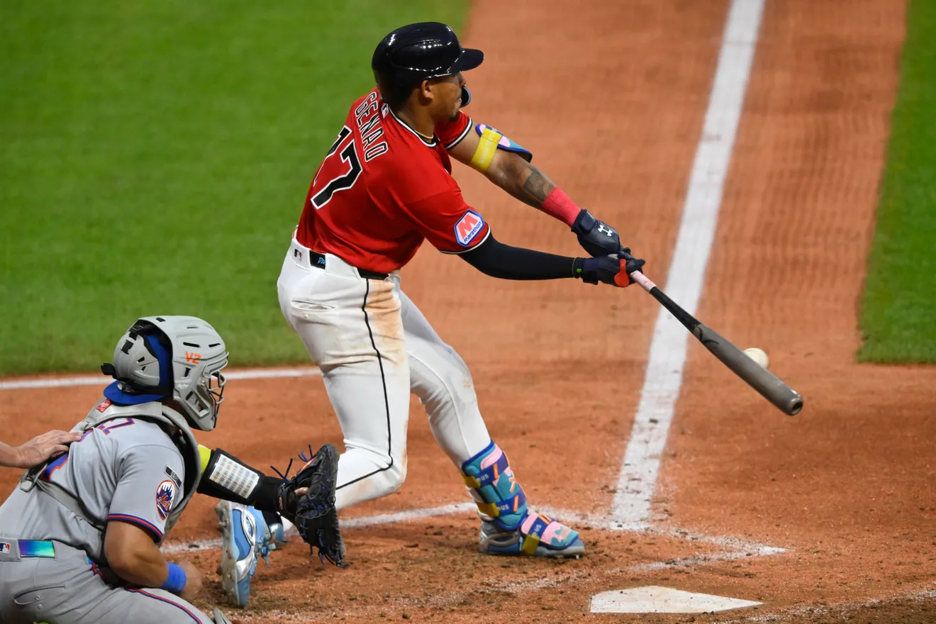 Baseball batter in a red jersey swings at a pitch near home plate with a catcher in gray gear behind him.