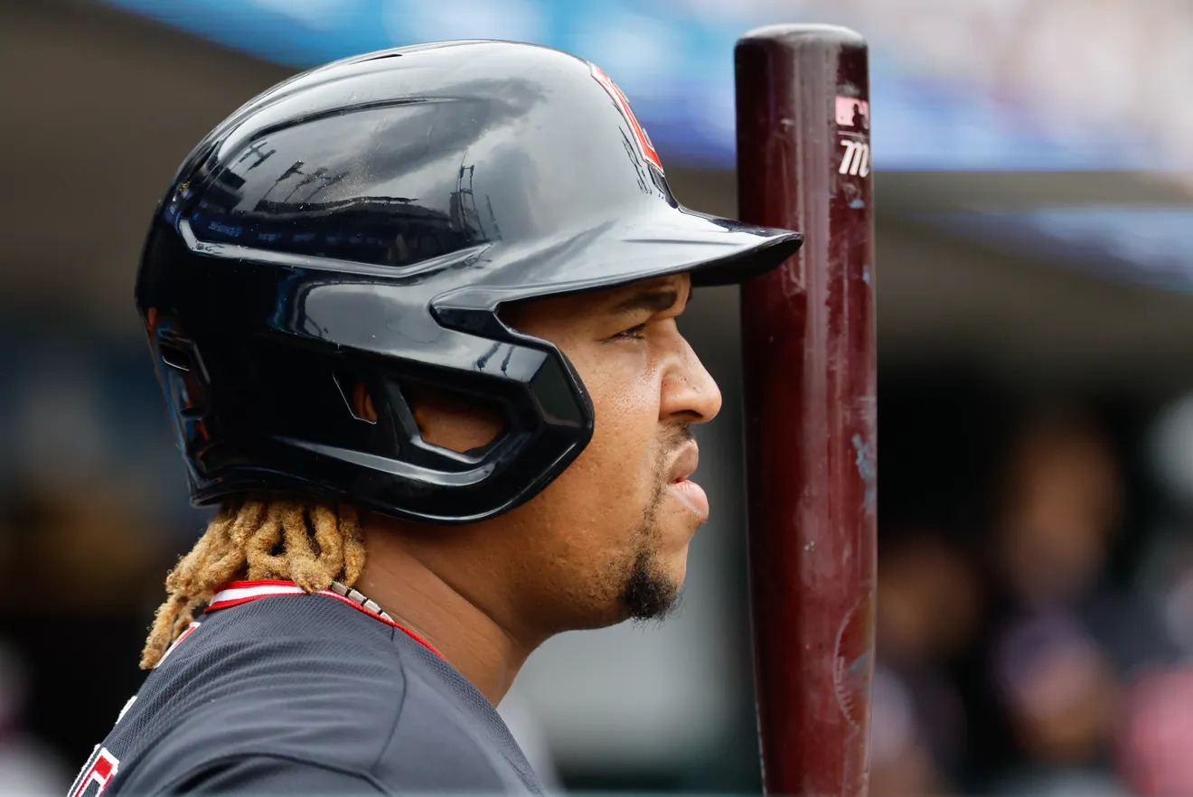 Baseball player in a black batting helmet and gray uniform, standing beside a wooden bat in the dugout.