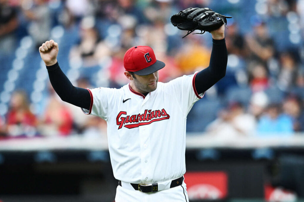 Cleveland Guardians pitcher in a white uniform and red cap raises his right fist while holding his glove above his head on the mound, crowd blurred in the background.