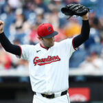 Cleveland Guardians pitcher in a white uniform and red cap raises his right fist while holding his glove above his head on the mound, crowd blurred in the background.