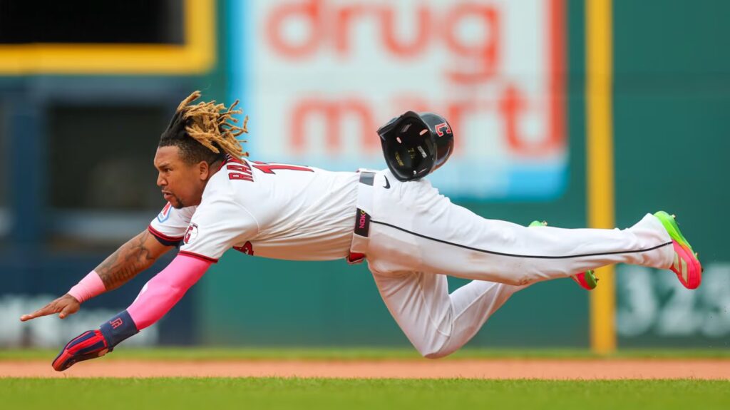 Baseball player in a white uniform dives full length, reaching with his left arm as the helmet flies off midair.