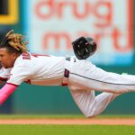 Baseball player in a white uniform dives full length, reaching with his left arm as the helmet flies off midair.