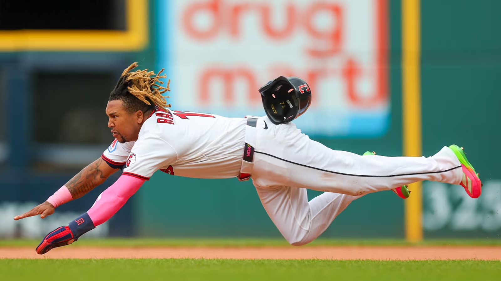 Baseball player in a white uniform dives full length, reaching with his left arm as the helmet flies off midair.